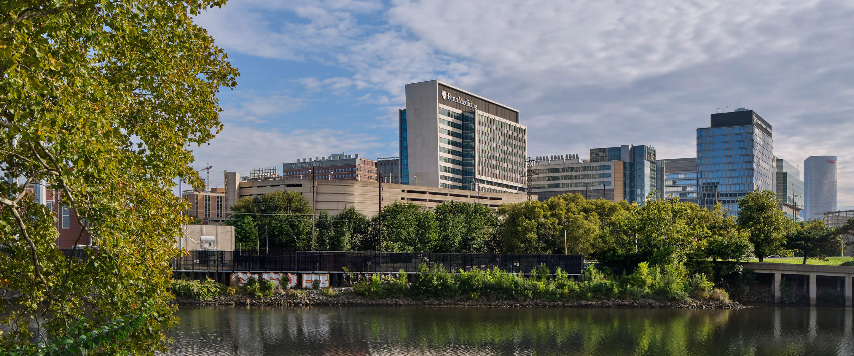 City skyline featuring various buildings, including Penn Medicine, alongside a river and lush greenery. Clear skies above.