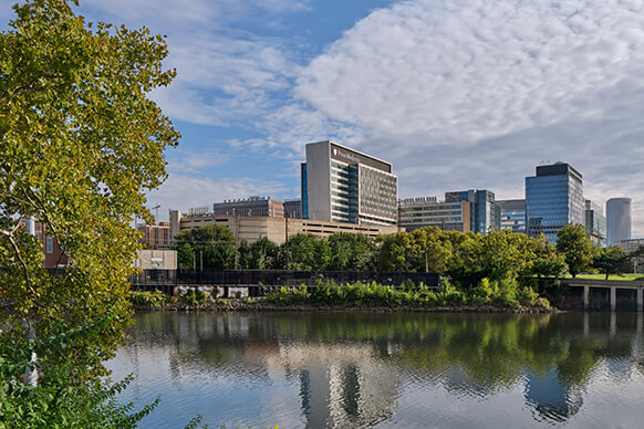 Cityscape featuring commercial buildings along a riverbank, surrounded by trees under a partly cloudy sky. Reflections visible on the water.