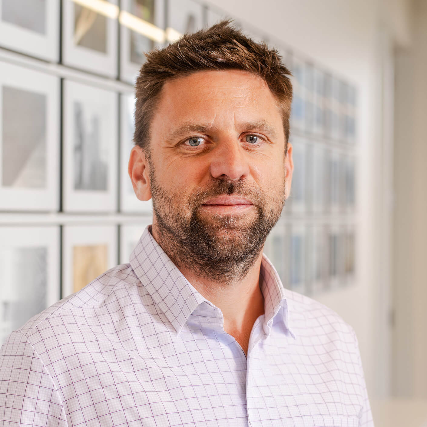 Portrait of a man with a beard, wearing a checked shirt, standing in front of a wall displaying framed photographs in an office setting.