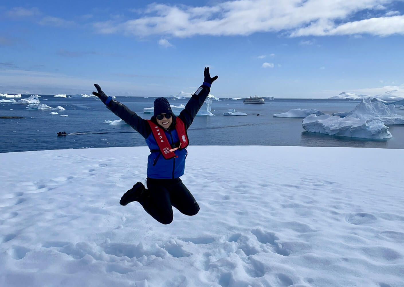 Person in a life jacket jumps joyfully on snow-covered ground with icebergs and a boat visible in the background. Clear blue sky overhead.