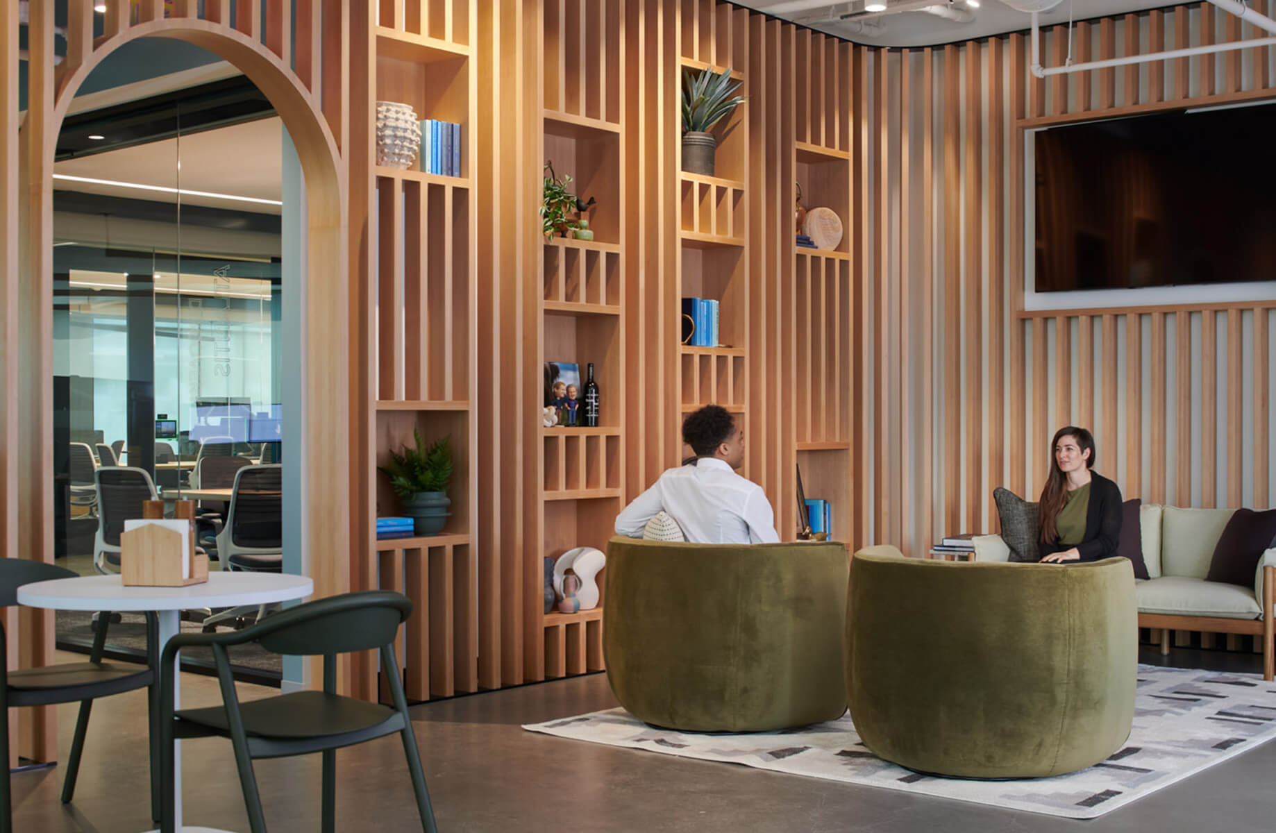 Two people seated in curved green chairs, discussing in a wood-accented lounge area featuring shelves and plants. An office space is visible in the background.