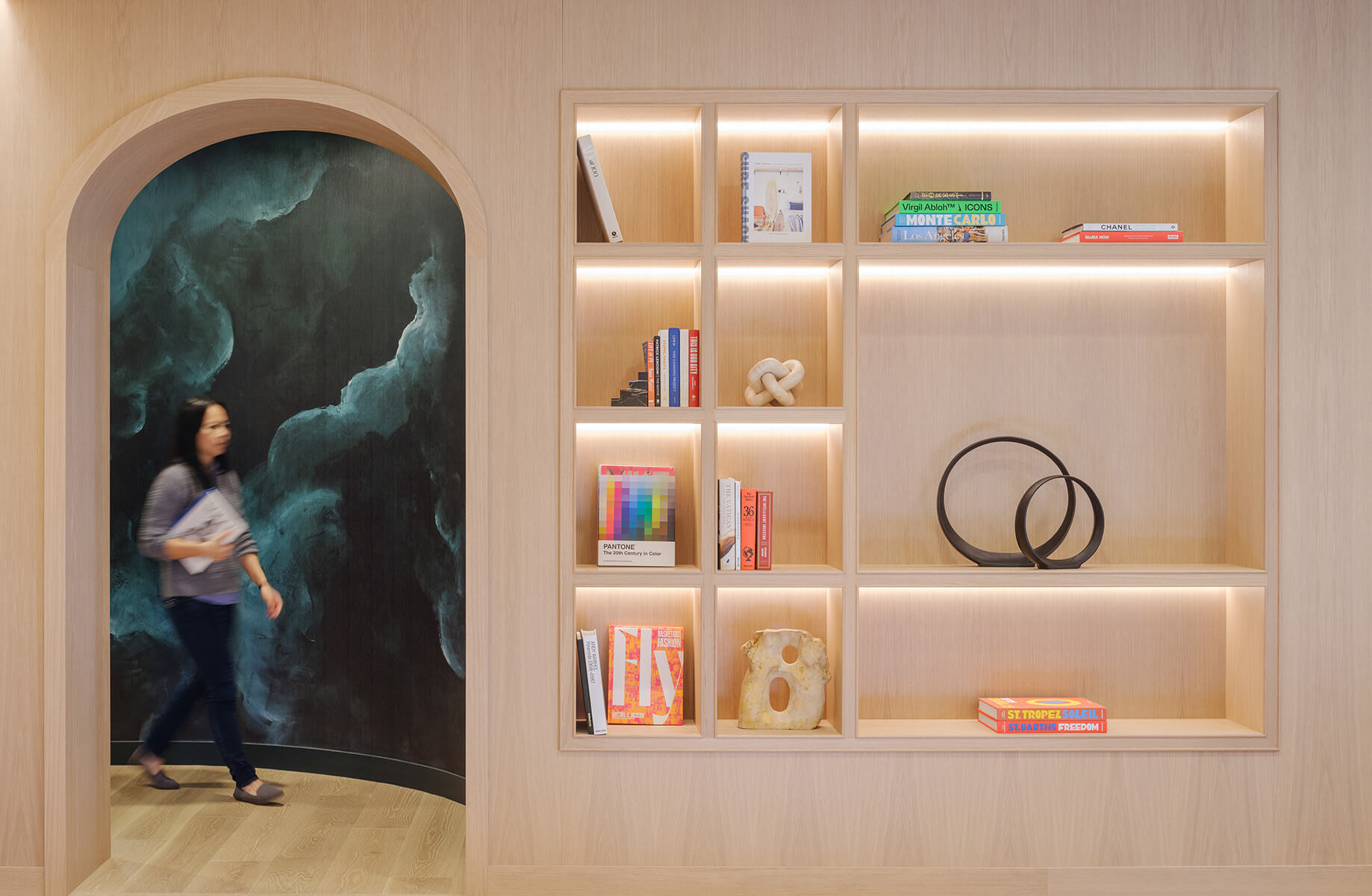 A woman walks past a bookshelf featuring various books and decorative items in a contemporary interior space. Soft lighting highlights the shelves.