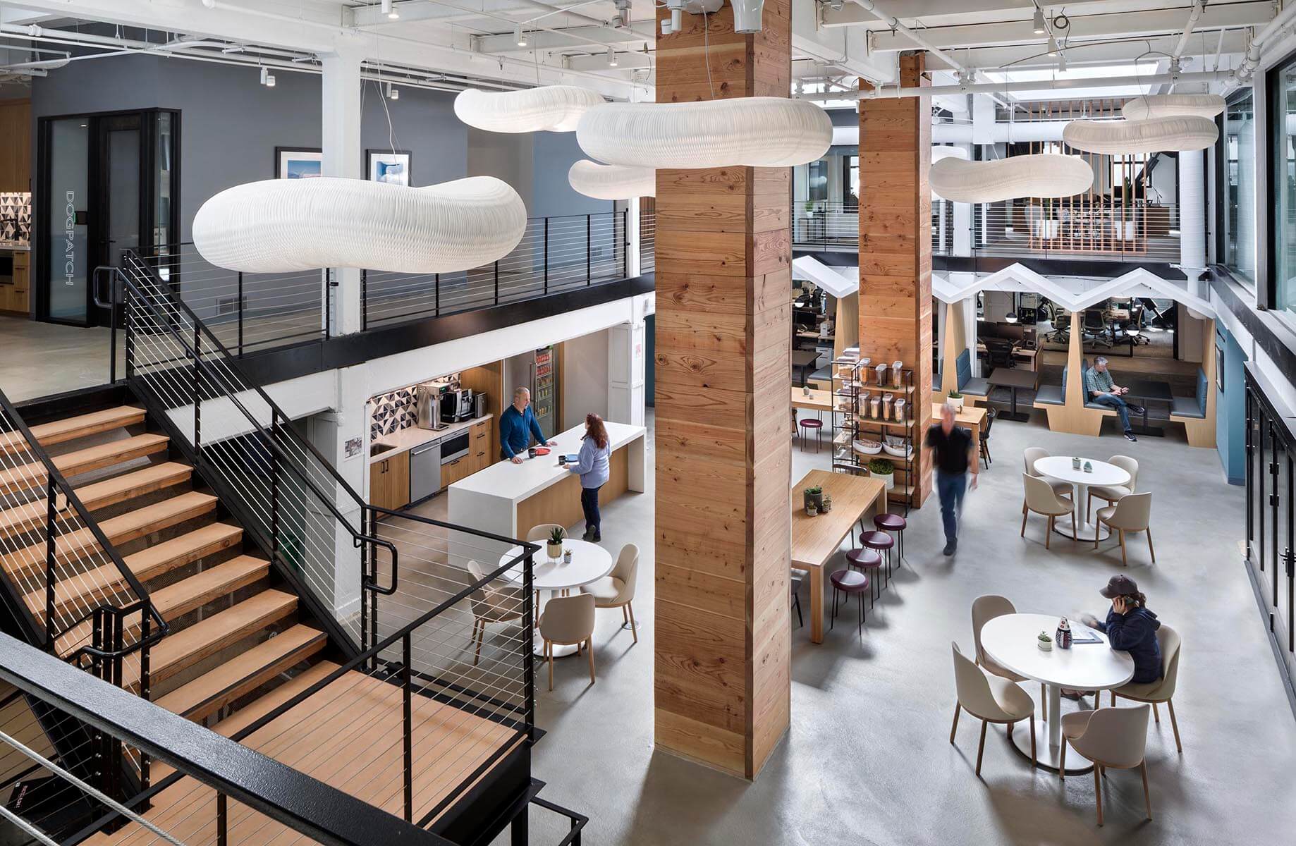 Open office interior featuring wooden pillars, sleek furniture, and a kitchen area with people engaging in casual work and conversation.