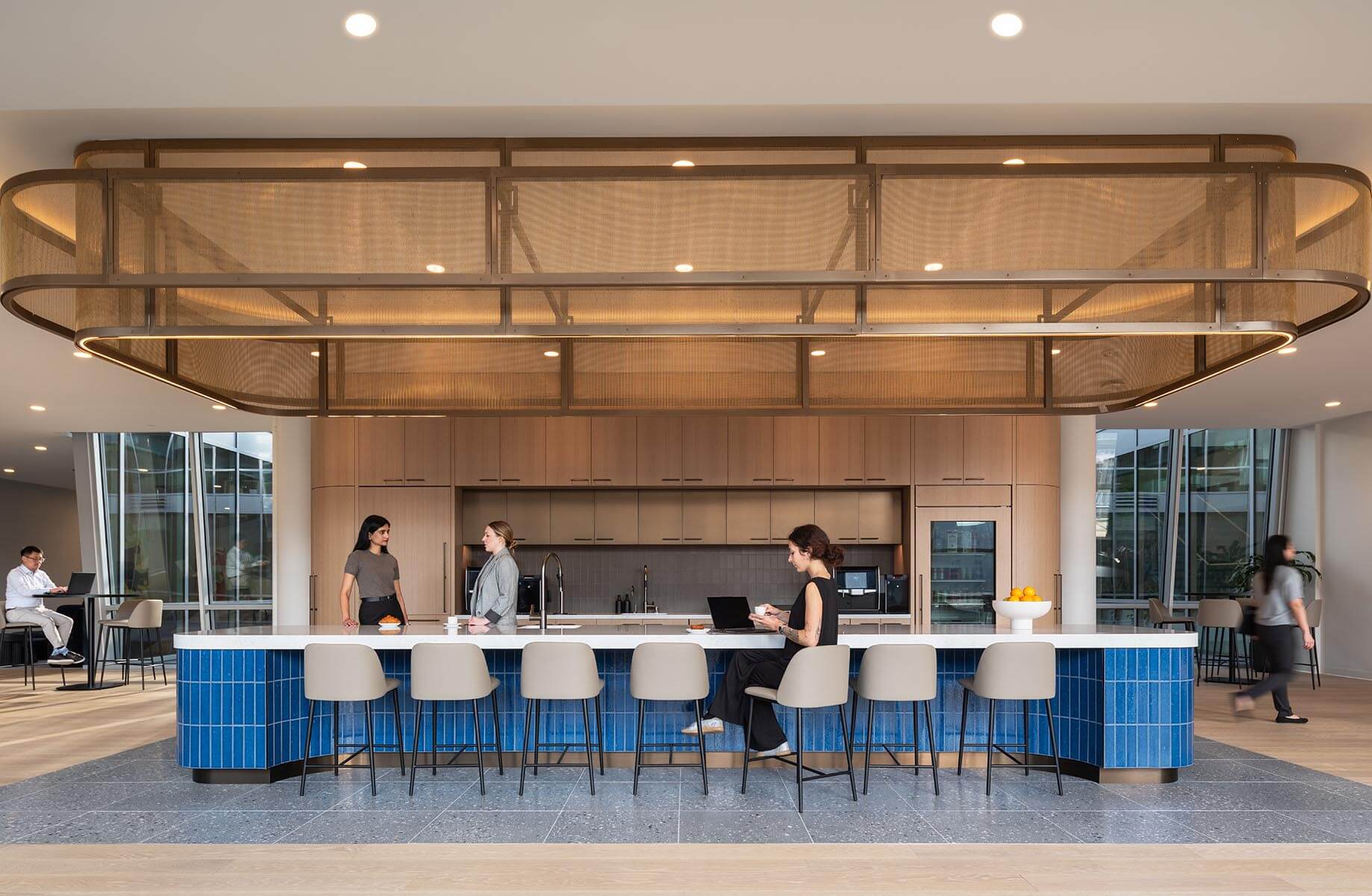 Contemporary office kitchen with blue tile island, seating, and staff interacting, featuring wood cabinetry and large windows.