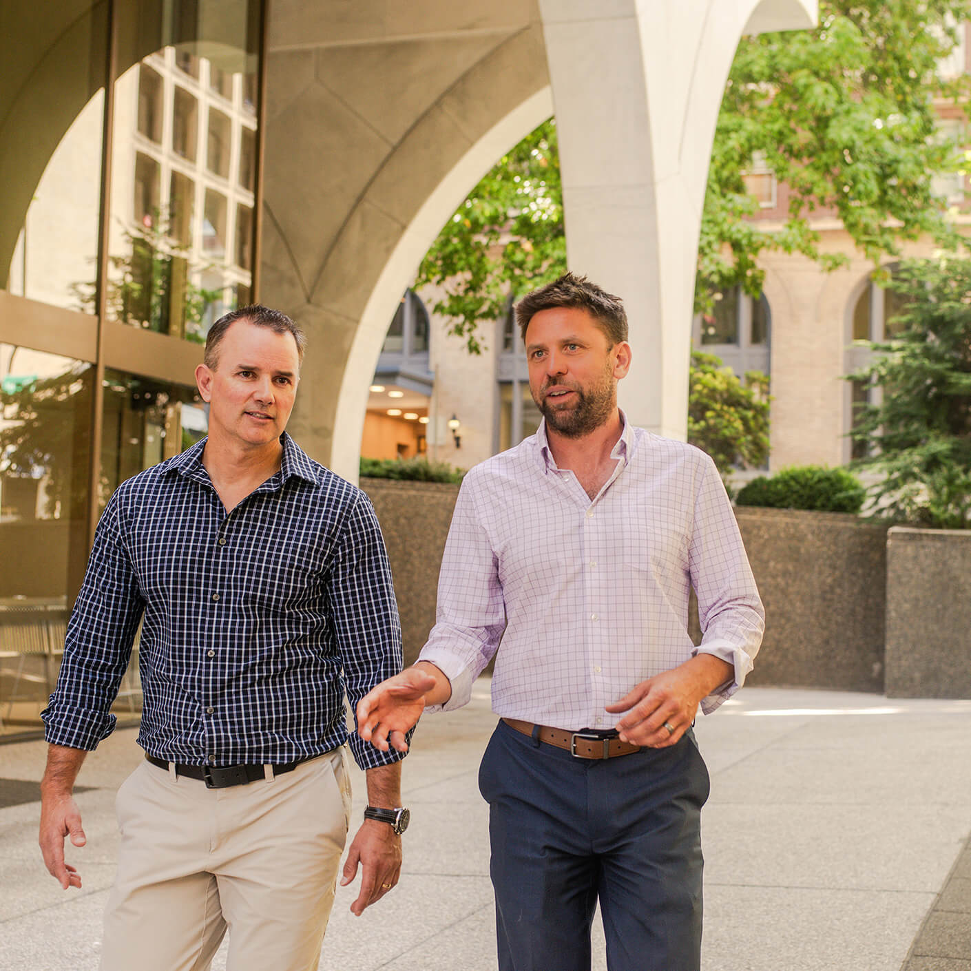 Two men in business attire walking outside near a large building, engaged in conversation under a concrete archway.