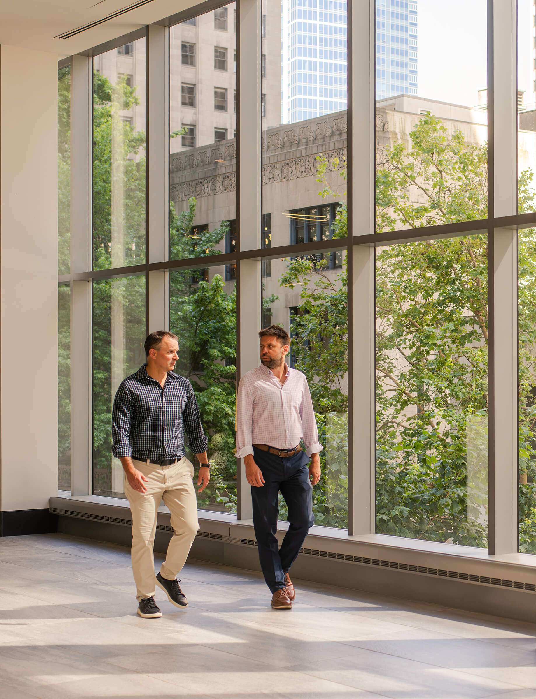 Two men walk side by side in a well-lit space with large windows and greenery visible outside, showcasing a professional setting.
