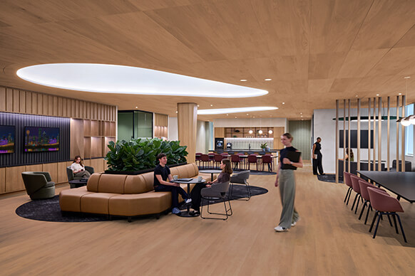 Office lounge area featuring wooden elements, seating arrangements, lush greenery, and a kitchenette with people engaged in various activities.