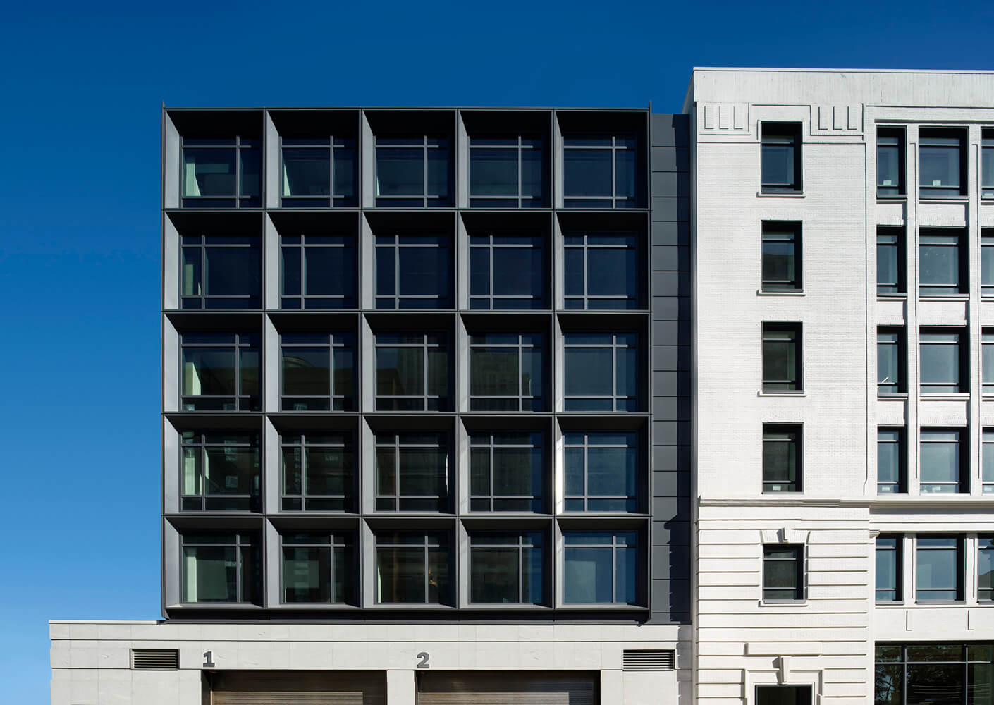 Facade of a building featuring a grid of windows on the left and a textured surface on the right, set against a clear blue sky.