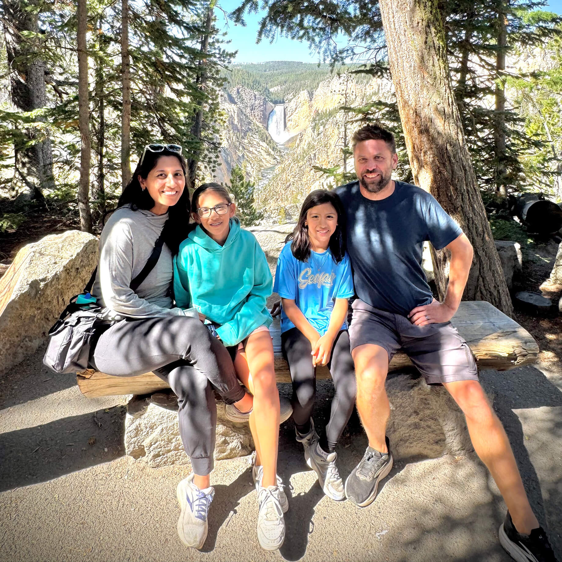 Family enjoying time together on a bench in a forest setting, with trees and a waterfall visible in the background.
