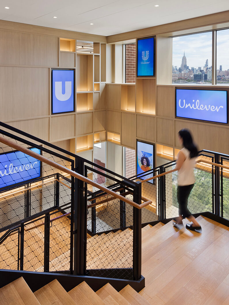 Staircase in Unilever office featuring wood accents, framed screens, and a view of the New York City skyline. Person walking down the stairs.