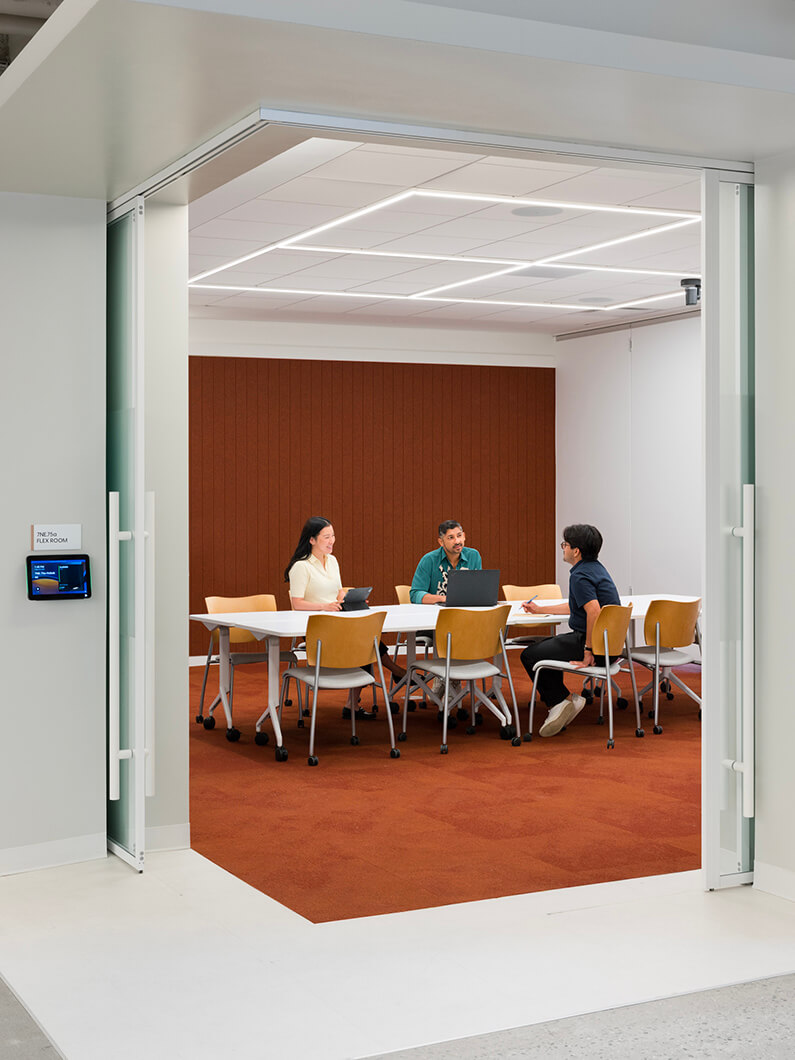 Four people engaged in a meeting around a table in a conference room with a warm-colored carpet and soft lighting.
