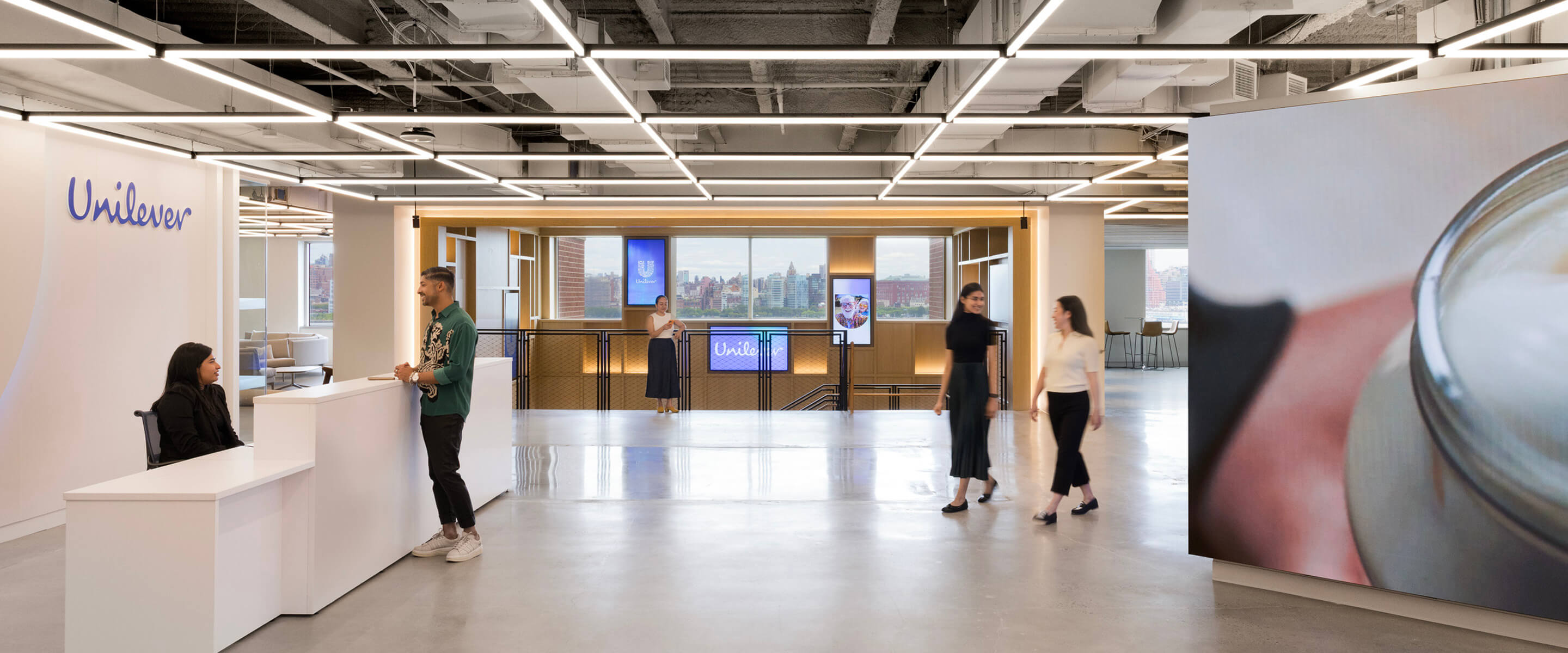 reception area at Unilever office with two employees at the desk, others walking, digital screens, and city view through large windows.