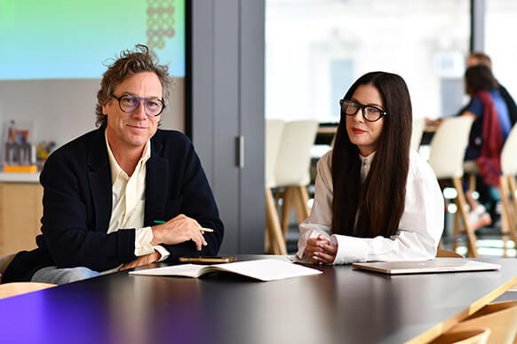 Two professionals sitting at a conference table, engaged in discussion, with notes and a laptop in a bright, open office environment. 